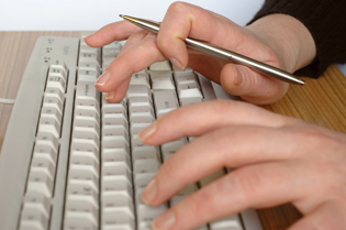 Hands typing on a keyboard, holding a pen